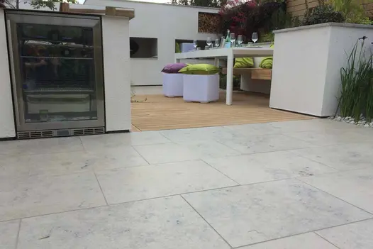 Low view across Jura Grey smooth limestone paving to decked eating area with table, cushions and inbuilt chiller unit. 