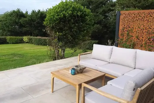 Corner sofa and table with metal screen behind sit on Jura Grey smooth limestone paving at edge of large lawn.