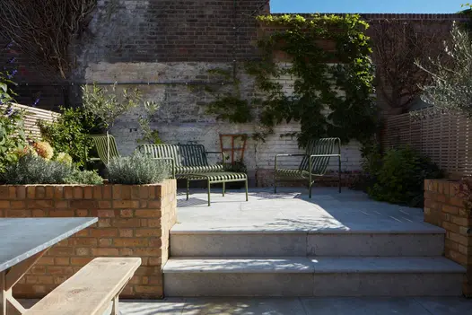 2 steps rise past brick raised bed to walled patio of Jura Grey sawn limestone paving with green metal furniture.