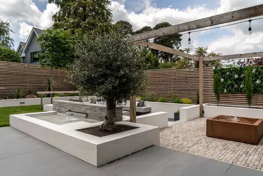 Water pours into pond from slot in low wall clad in Kandla Grey sandstone into pond on patio of mixed paving with sunken seating.