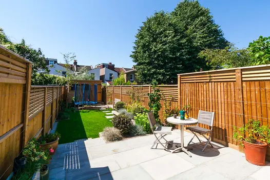 Long, narrow, fenced back garden, with Kandla Grey Indian sandstone patio, lawn with stepping stone slabs, and trampoline at end.