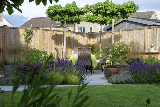 Garden patio of Kandla Grey Indian sandstone slabs, under leafy canopy, with rattan dining set surrounded by colourful planting.