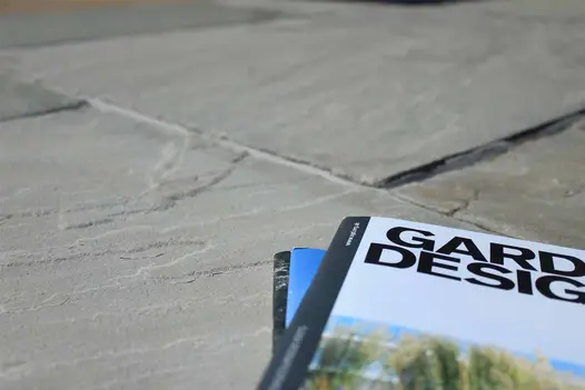 Close-up of Kandla Grey Indian sandstone paving, showing riven surface texture, with 2 Garden Design journals to right.