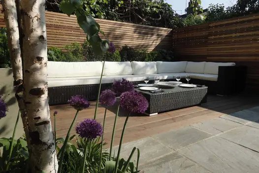 Long outdoor sofa on decking that abuts Kandla Grey Indian sandstone paving laid stack bond. Cedar fence on 2 sides. 