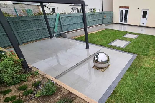 Silver ball sits on gravel square on 2-level patio of Kandla Grey porcelain, with dark plank edging in back garden with lawn.
