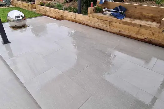 Large silver ball at end of 2-level area paved in Kandla Grey porcelain slabs, next to sleeper retaining wall with inbuilt bench.