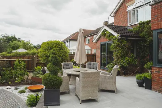 Light rattan furniture and dark planters sit on Kandla Grey Porcelain Paving, ivy clad back porch with french doors in the distance.