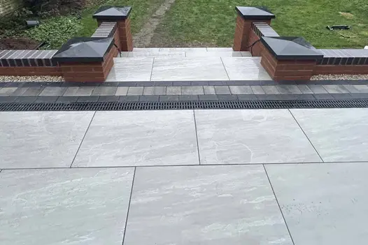 3 steps descend between brick walls and pillars with black pier caps from Kandla Grey porcelain paving with channel drain.