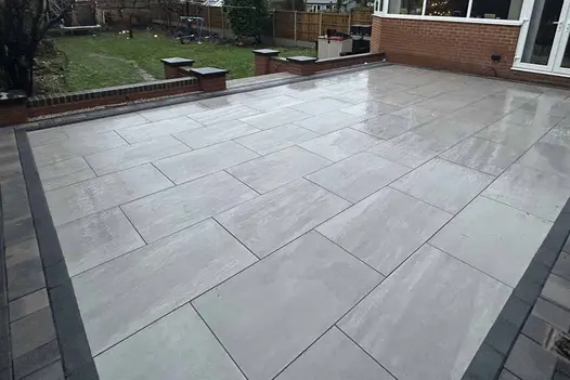 At side of house french doors open onto patio of Kandla grey porcelain paving with steps down to large grassed garden.