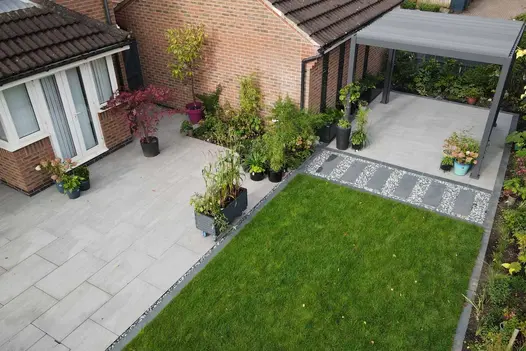 Two patios of Kandla Grey porcelain paving displaying pots filled with plants, broken up by lawn and pergola.