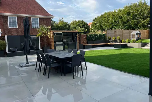 Metal dining set and furled parasol on large wet patio of Yard 1200x1200 porcelain paving. Outdoor kitchen in background.