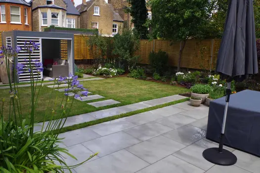 Contemporary Grey sawn sandstone stepping stones cross grass from house to matching paved area with freestanding parasol.