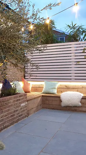 Corner of Kota Blue limestone patio with brick raised bed and L-shaped wooden bench with cushions under slatted fence.