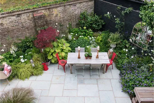 Kota Blue limestone patio edged on 2 sides with deep varied planting and tall brick wall. 2 candlesticks on wooden outdoor table.