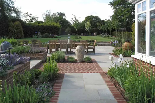 Large patio of Kota Brown limestone slabs and clay pavers, with brick raised beds, pithoi water feature, and 3 steps up to lawn.
