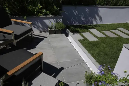 Plank-shaped paving slabs lead across lawn next to wall to Flamed Grey sawn sandstone patio with 2 chairs. Built by Landform.