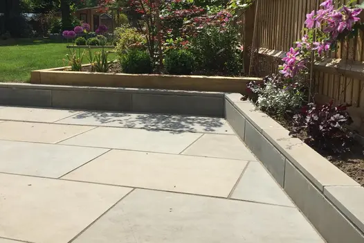 Oblique-angled corner of patio of Heath smooth sandstone paving, with raised bed edging of matching slabs cut to size.
