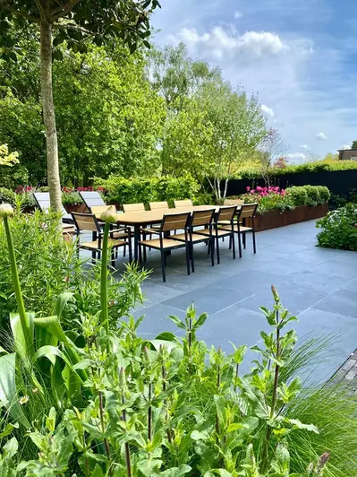 View across tall leafy plants to 10-seater dining table on large area pavied with Brazilian Black slate slabs. Design by Greencube.