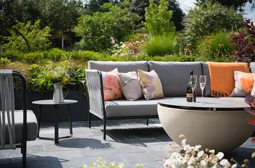 Grey-cushioned garden sofa and table on Brazilian Black stone slabs with lush planted beds in large garden behind.