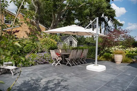 Large patio of Brazilian Black slate slabs with wooden dining set under cantilevered parasol, in midst of flowering beds.