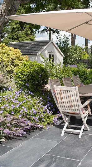 Flowering border spills over onto Brazilian black slate patio with wooden dining set and parasol. Garden shed in background.