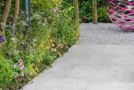 Silver-grey porcelain paving leading to white gravel with spherical pink and silver metal sculpture, edged by lush flower border.