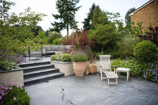 Tall bushes and trees enclose brazilian black slate patio with bullnose steps leading up to outdoor lounge area.