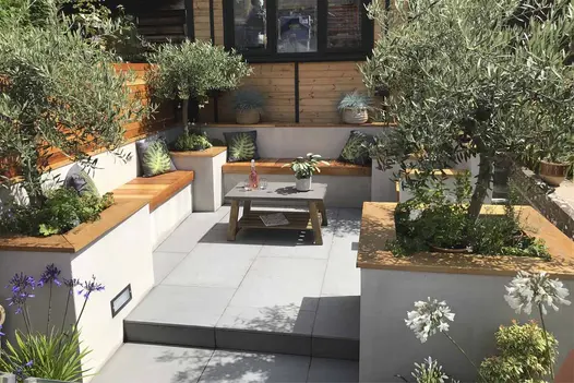 Small back garden paved with Trendy Black porcelain, lined with white, wooden-topped benches and raised beds with olive trees.