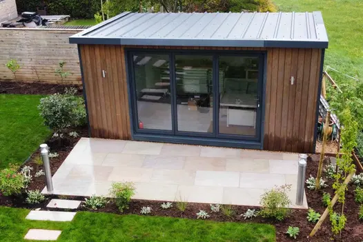 Garden building encased by freshly dug flowerbeds with small patio of raj green porcelain paving as the entrance.
