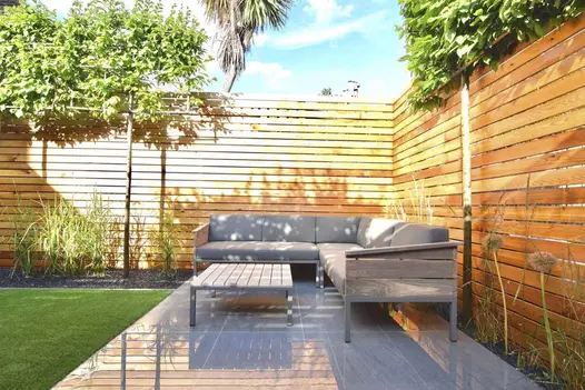 Corner of fenced garden lined with box-head hornbeams. Corner lounge set sits on Trendy Black Porcelain paving by artificial lawn.