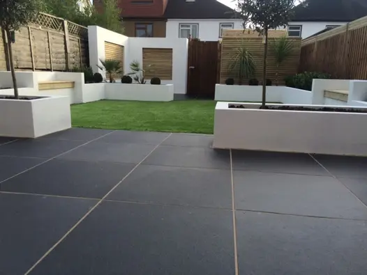 White-painted raised beds edge colour- enhanced Midnight Black limestone paving with pale joints in modern garden with high fence.