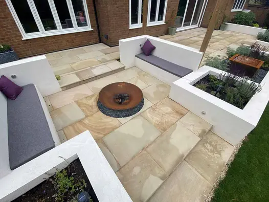 2 inbuilt benches face each other across Corten steel firebowl on wide patio of Light Buff smooth sandstone at back of brick house.