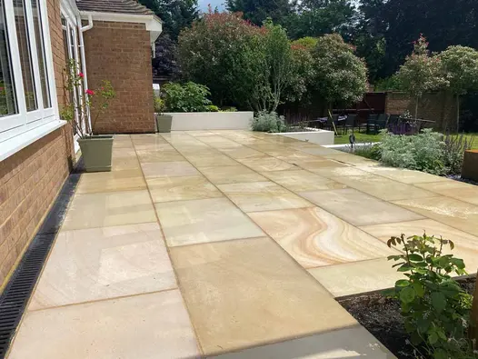 Wide patio of Light Buff sawn sandstone paving slabs runs across back of house, with lawn on lower level to right.