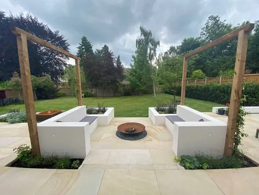 2 identical constructed benches of linear design face each other across fire bowl on patio of Light Buff sawn sandstone paving.