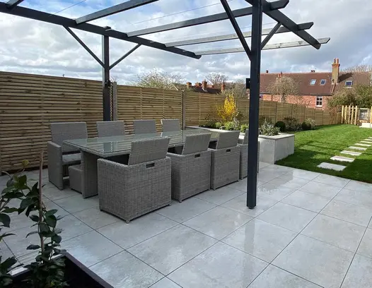 Low maintenance family dining area on top of a Porcelain patio with a metal pergola above and cladded raised beds.