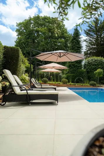 Low level view of light grey porcelain paving used around swimming pool, six sunloungers sit under 3 sun shades. 