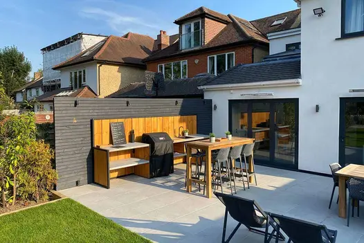 Patio of Florence Grey porcelain paving with 6-seat bar table by outdoor kitchen against fence. Design by Limebok Landscaping.