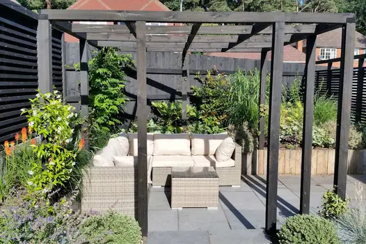 Wooden pergola over rattan L-shaped sofa and table in corner of garden with raised beds and Midnight Black limestone paving.