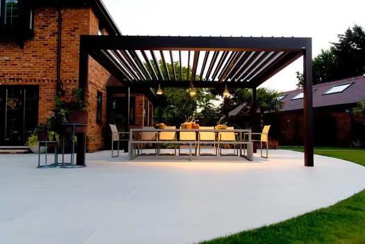 Huge louvred pergola over 12-seat outdoor dining set on large Faro porcelain paving patio that curves around corner of brick house.