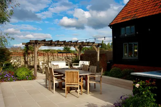 Slab Khaki Porcelain Paving used in outside dining area. Pergola sits behind wooden table with 7 seats surrounding.