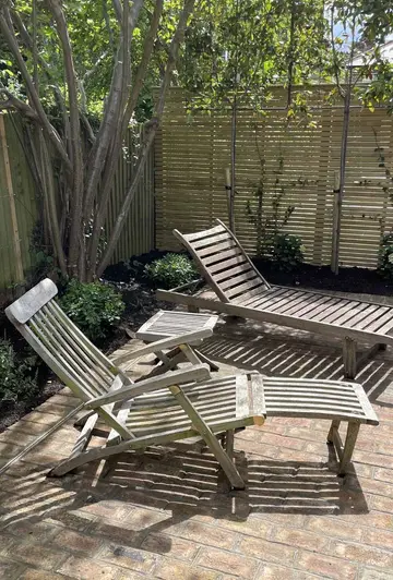 Two sunloungers casting shadows on london mixture clay paver patio with fence and tree in the background.
