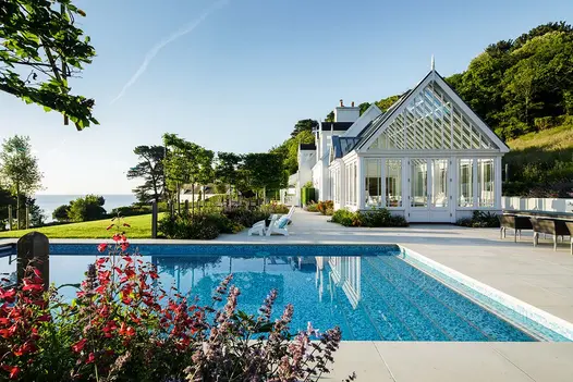Large swimming pool with Beige smooth sandstone paving surround and view over lawn to the horizon. Design by The Lovely Garden.