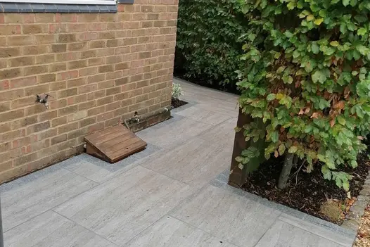 Small Travertine Dark porcelain patio edged with sawn granite setts and bordered by deciduous hedging plants.