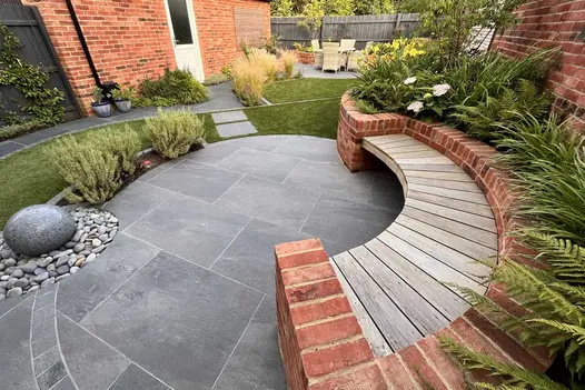 Oval patio of Platinum Grey porcelain paving, part edged with curved raised bed with inset wooden bench. Design by Mark Langford.