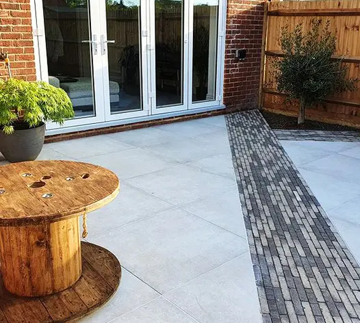 Diagonal stripe of Silver Multi Clay pavers runs to corner of house past giant bobbin table on grey paving by Marli Construction.