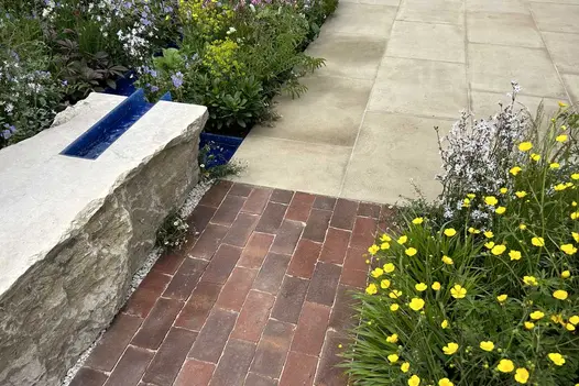 Clay paver path between low wall and yellow-flowered plant meets Britannia Buff sawn Yorkstone paving.
