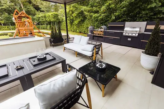 Covered patio of Faro porcelain slabs, with outdoor kitchen against hedge on far side and play fort on lawn to left.