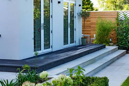 Two steps lead up to narrow decking area on porch of french windows, the burnt cedar colour stands out against the white of the house.