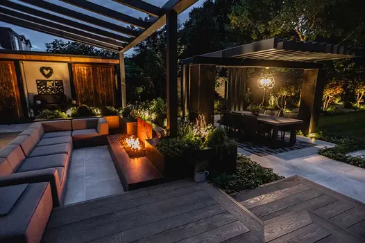 Corner sofa under pergola at night sits on golden oak millboard decking, paved area in the distance with dining table.