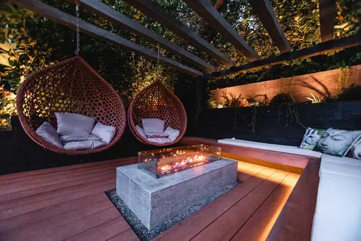 Two egg chairs float from wooden pergola in cosy firepit are with corner bench on coppered oak millboard decking.